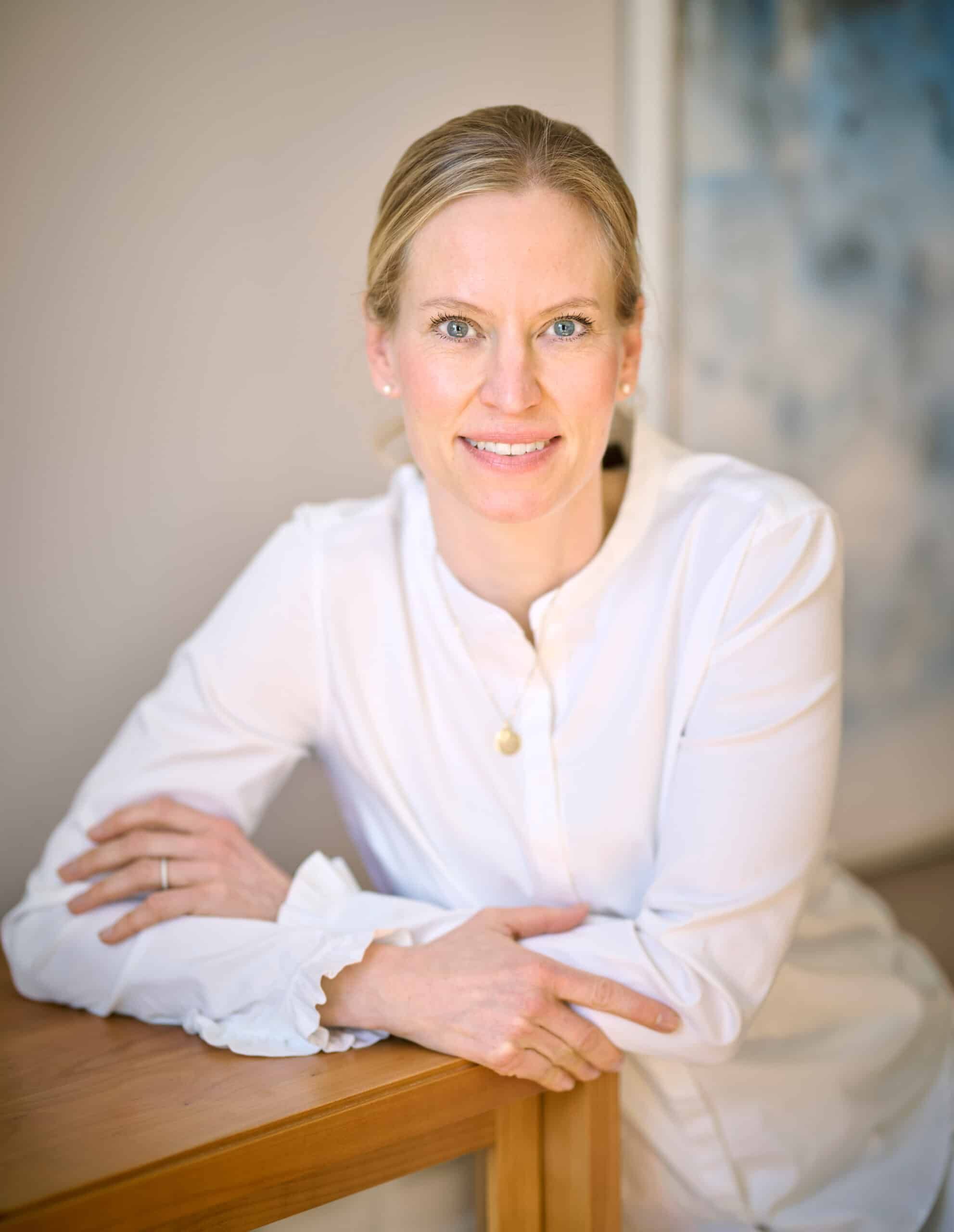 Dr. med. Ursula Orth-Tannenberg, eine lächelnde Frau mit blondem Haar in weißer Bluse, sitzt in einem hellen Raum und blickt freundlich in die Kamera.“