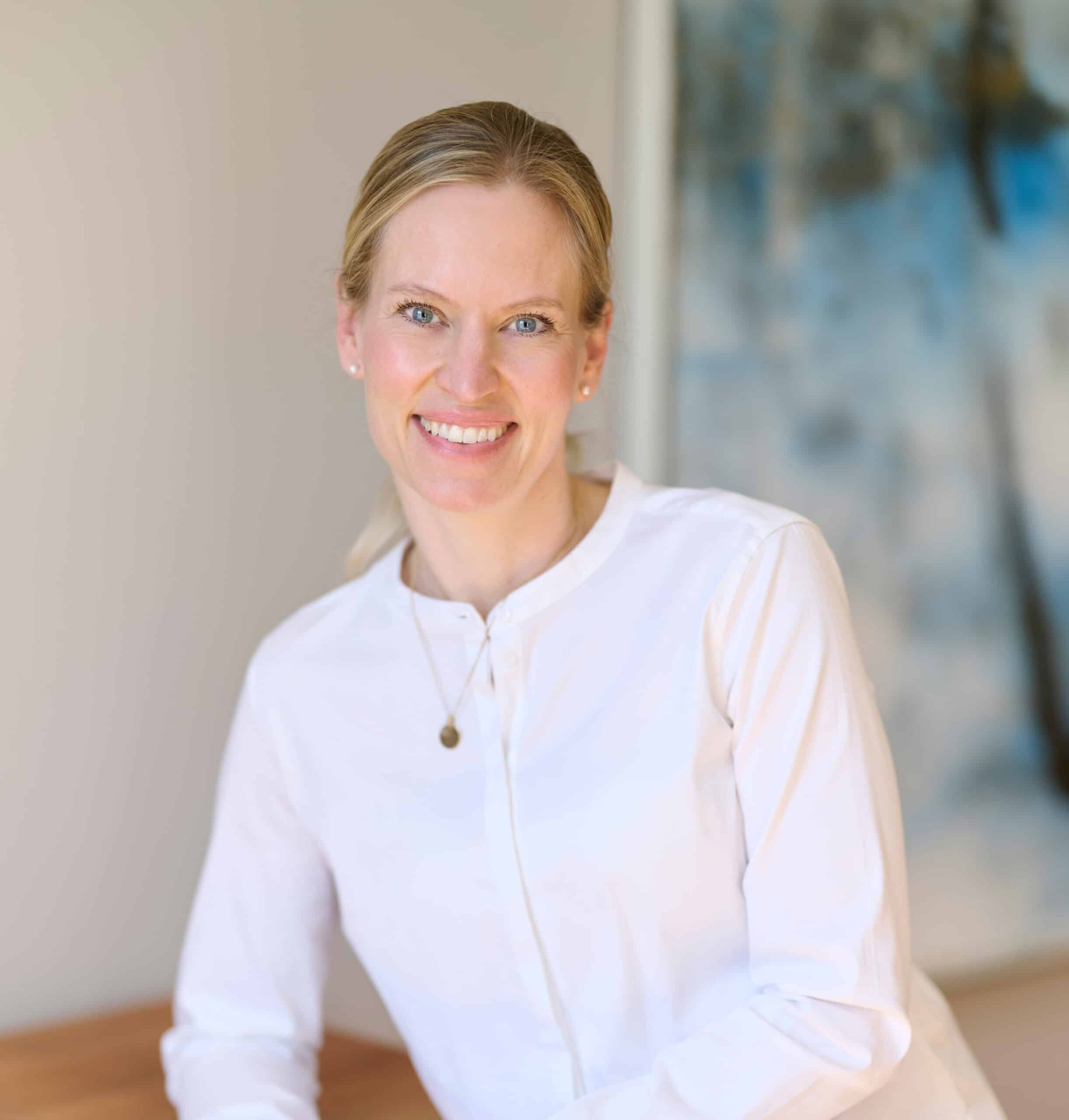 Dr. Ursula Orth-Tannenberg, eine Frau mit blondem Haar in weißer Bluse, lehnt sich freundlich lächelnd auf einen Holztisch.